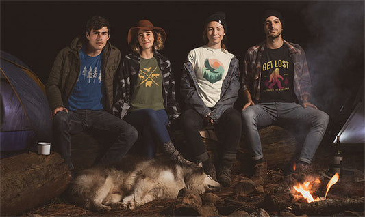 Friends gathered around a campfire at night, wearing outdoor-themed graphic t-shirts, with a camping tent and a dog resting nearby.